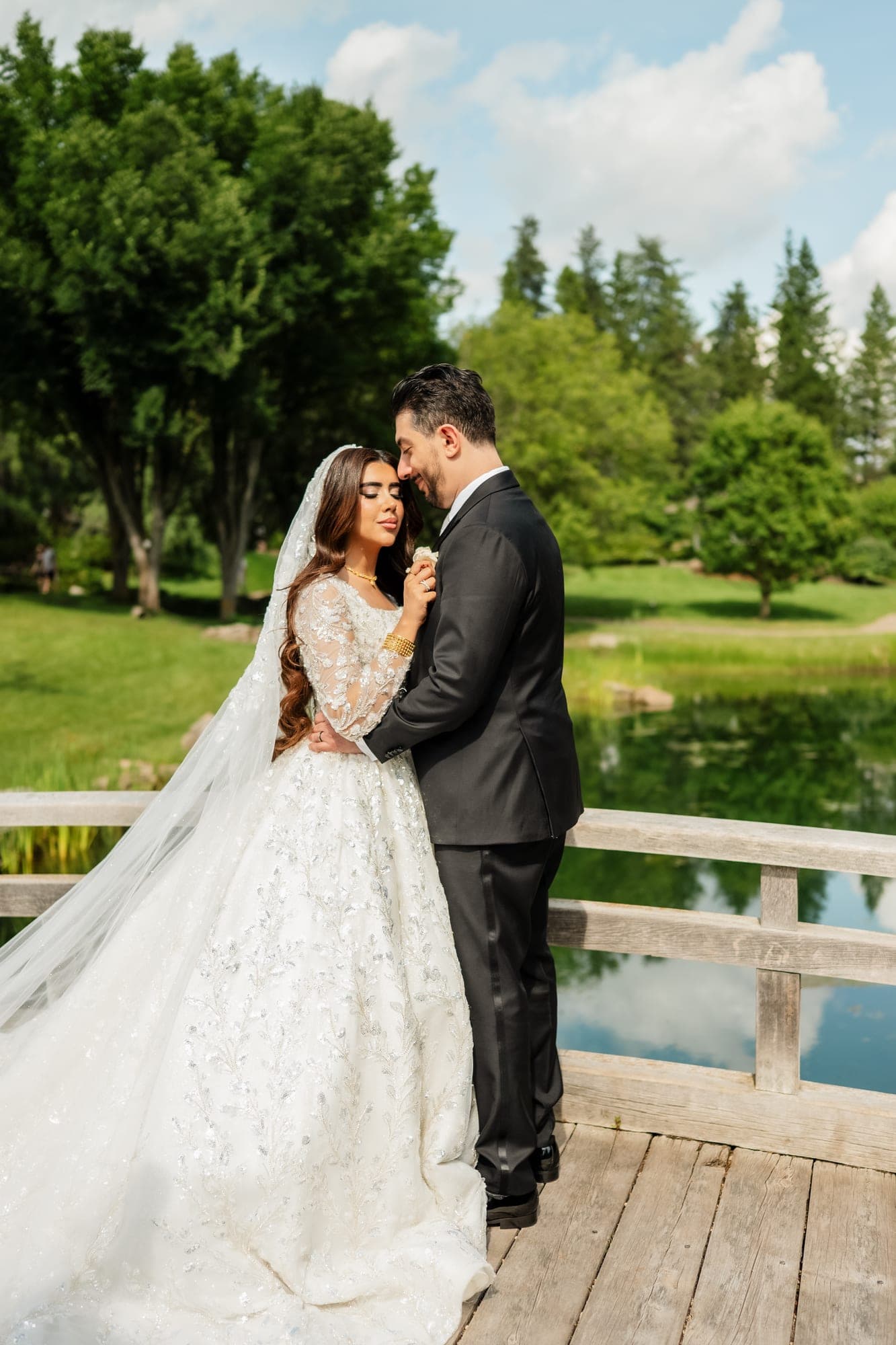 A bride and groom embracing on a wooden bridge beside a green garden pond